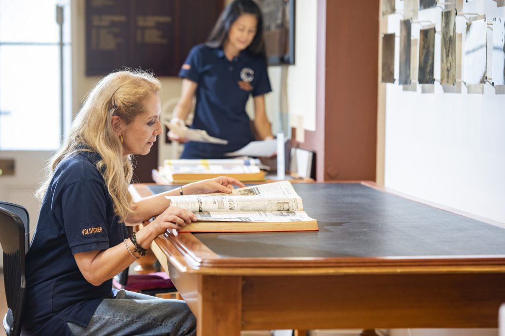 Volunteers reading historical materials at a long table beneath a wall of photographs